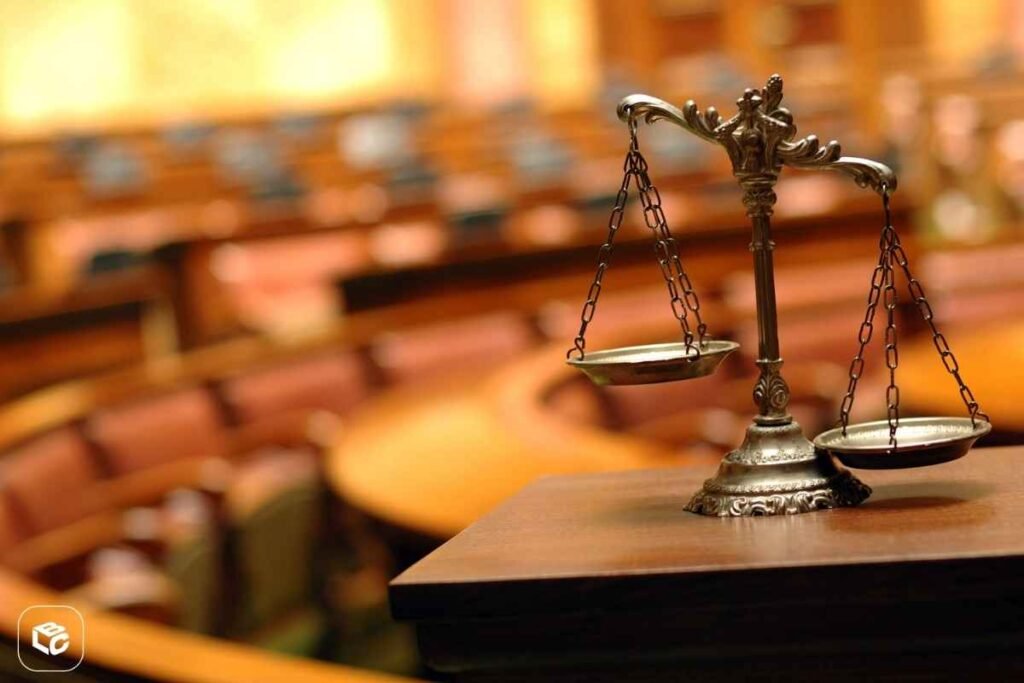 : Balança da Justiça em metal posicionada sobre uma mesa de madeira, em destaque no primeiro plano, com profundidade de campo reduzida. Ao fundo, um ambiente semelhante a tribunal ou sala de audiências aparece desfocado, com mesas e assentos em tons amadeirados. A imagem simboliza justiça, processos judiciais, direito e decisões legais. Logotipo da empresa visível no canto inferior esquerdo.