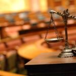 : Balança da Justiça em metal posicionada sobre uma mesa de madeira, em destaque no primeiro plano, com profundidade de campo reduzida. Ao fundo, um ambiente semelhante a tribunal ou sala de audiências aparece desfocado, com mesas e assentos em tons amadeirados. A imagem simboliza justiça, processos judiciais, direito e decisões legais. Logotipo da empresa visível no canto inferior esquerdo.